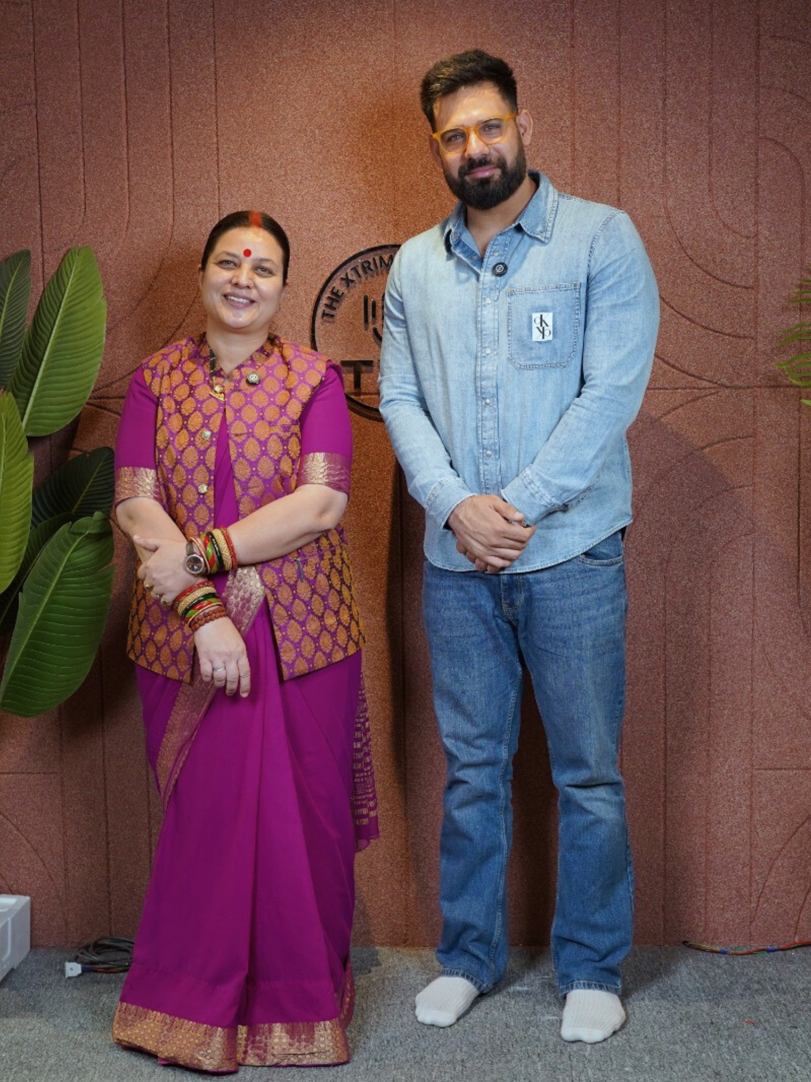 Woman in a purple saree with gold blouse and bangles standing next to a man in a blue denim shirt and jeans, posing together in a podcast studio with plants and "The Xtrim Podcast" logo in the background.