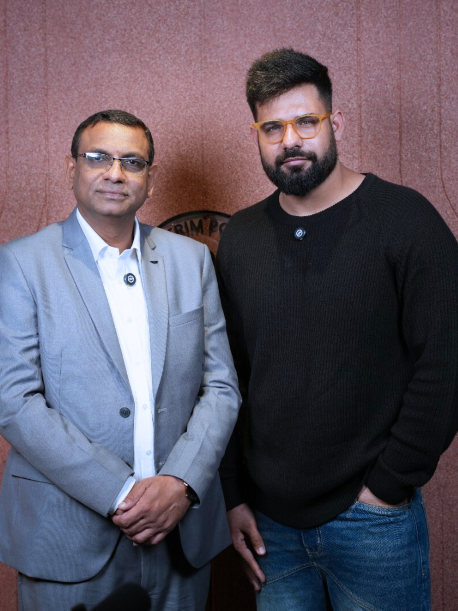 Two men standing in front of a podcast studio wall, one in a light gray suit and the other in a black sweater and blue jeans, both smiling for the camera.