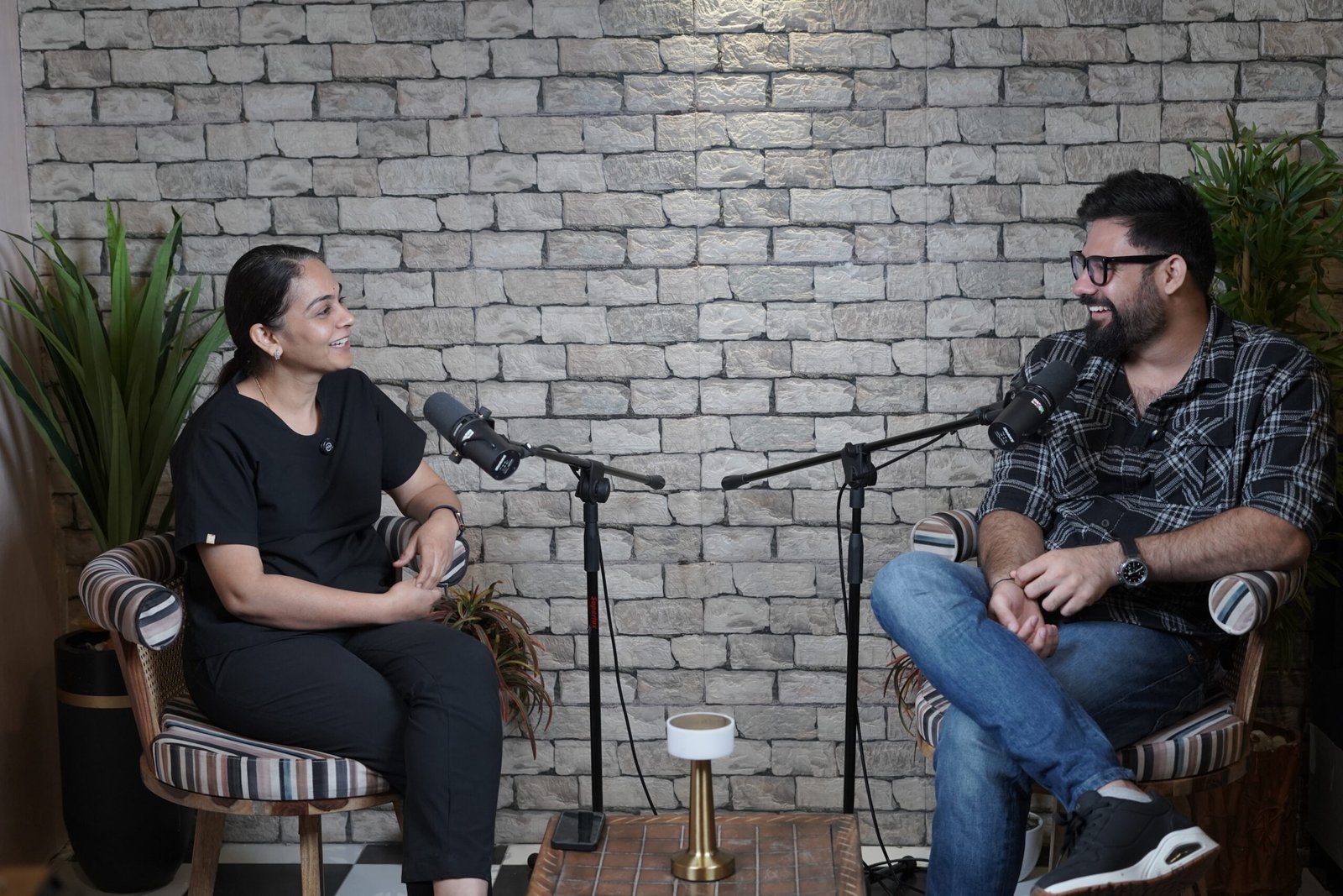 Woman and man seated in striped armchairs with microphones, smiling and conversing in front of a light gray brick wall, with potted plants behind them.