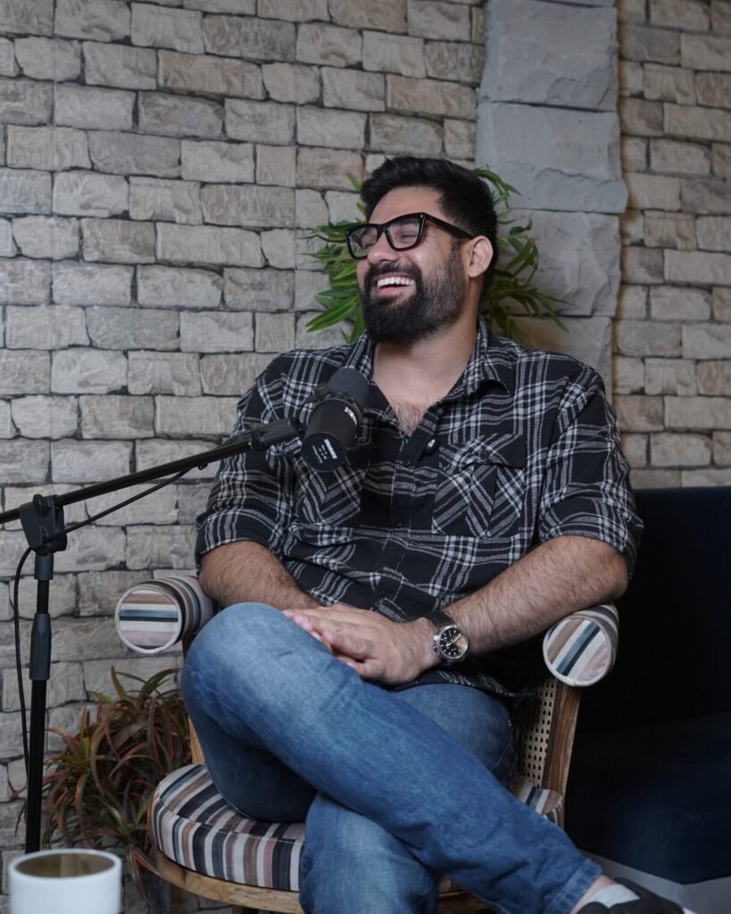 Podcast recording studio with two men seated under softbox lights, microphones and cameras set up, and a crew member standing on the right, with modern wall décor, shelves, and plants in the background.