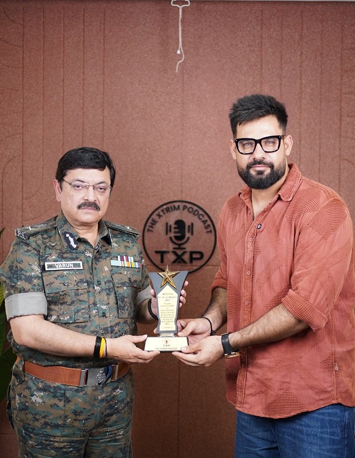 Two men standing in front of a brown podcast studio backdrop with "THE XTRIM PODCAST" logo, one in an army uniform and the other in a rust-orange shirt, holding a star-shaped award trophy together.
