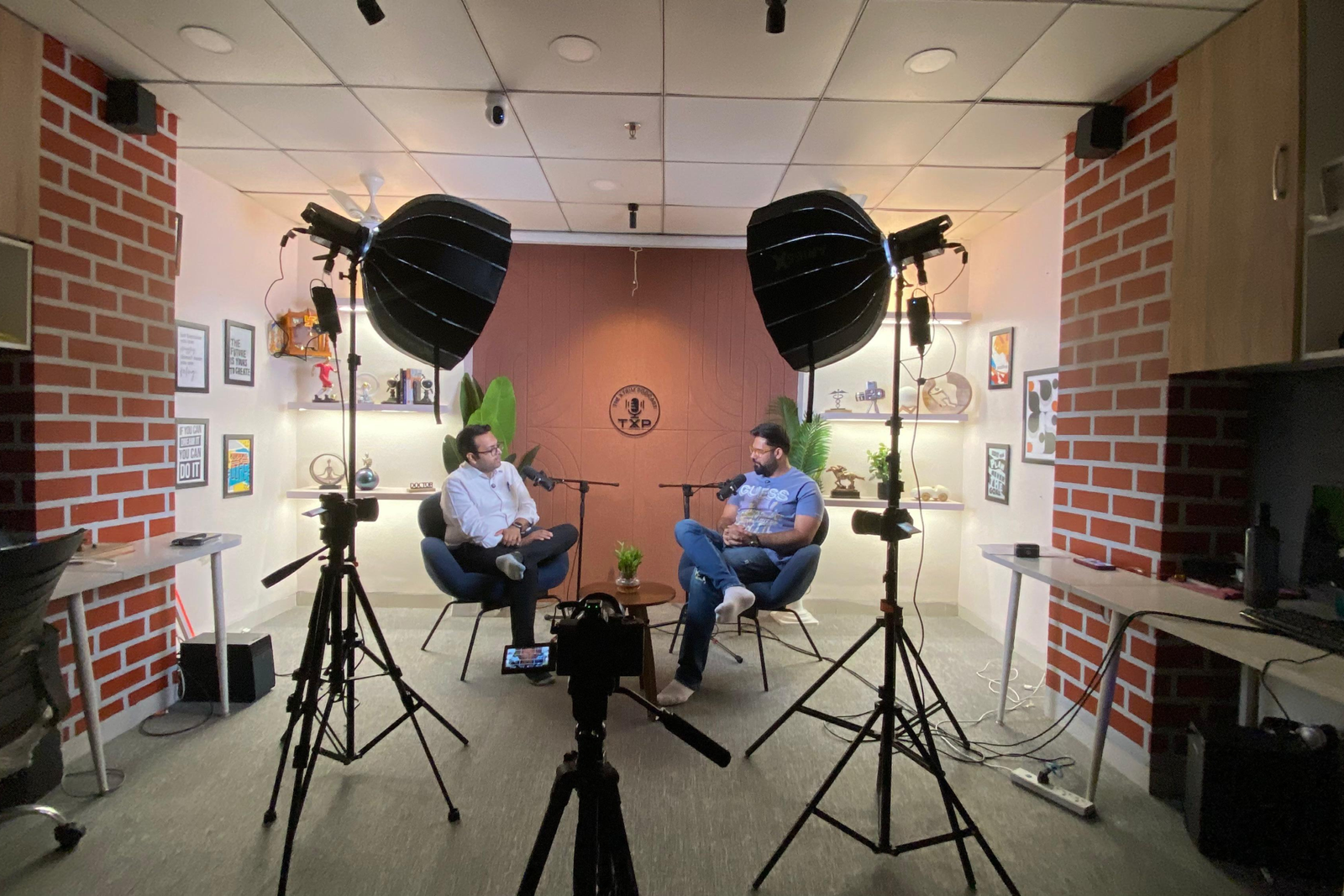 Podcast studio with two men seated in armchairs under professional lights, microphones between them, surrounded by cameras, decorated shelves, brick walls, and indoor plants.