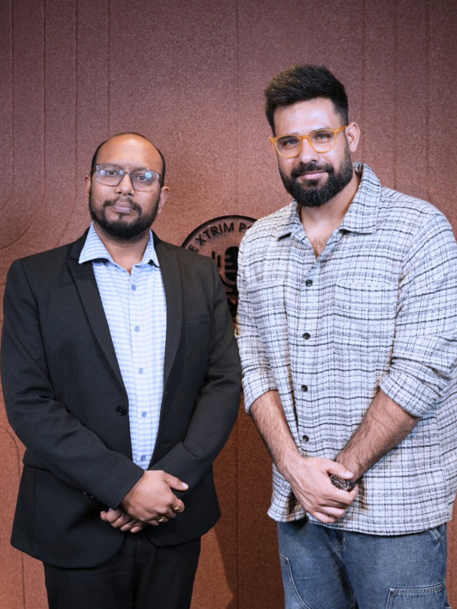 Two men standing in front of a brown wall with "The Xtrim Podcast" logo—one in a black suit and blue checked shirt, the other in a light checked shirt and jeans, both smiling and posing side by side.