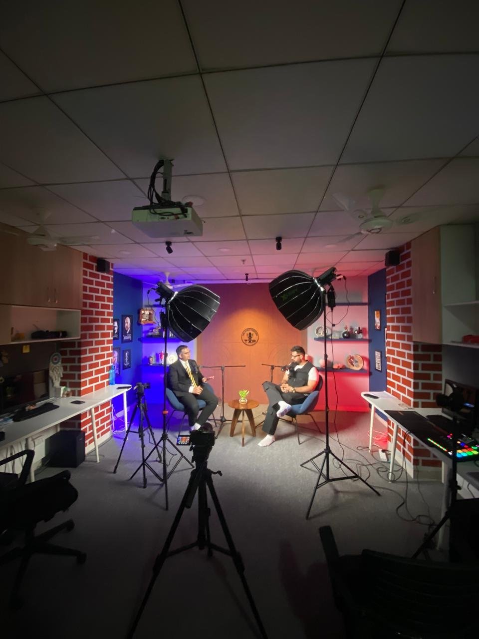 Woman in a green and white saree speaking with a man in jeans and patterned shirt, both seated in blue armchairs, microphones in front of them, recording a podcast in a modern studio with plants and decorative shelves.